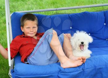 A blond smiling boy and a yawning puppy relaxing on a porch swing