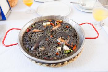 Paella negra on the pan, white table and glasses with juice
