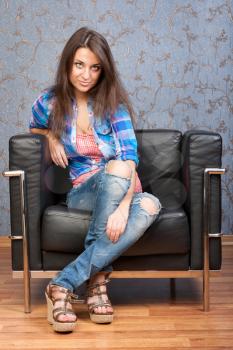 Portrait of beautiful brunette girl in a plaid shirt, sitting in leather chair against the wall with a retro pattern