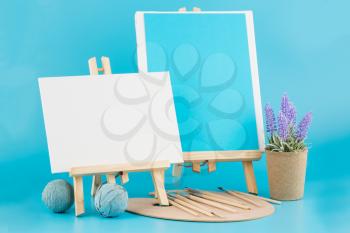 Two wooden easels with blank canvases, brushes and lavender flowers on blue background.