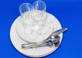 Pile of white plates, glasses with forks and spoons on silk napkin. Blue background