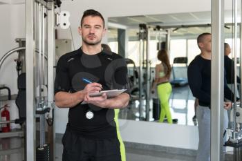 Personal Trainer With Clipboard Monitors People While They Exercise In A Gym Or Fitness Club