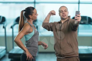 Bald Guy Showing Prety Young Woman How To Take A Selfie With A Cellphone In Fitness Center - Gym In The Background