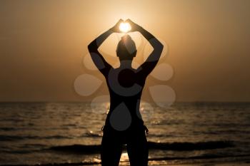 Beautiful Female Model Enjoying Sunset and Making Heart Sign on Sun at Seaside - Calm Water Reflects Silhouette of Woman - Sun Goes Under Horizon