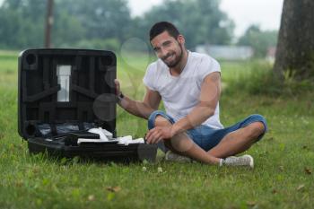 Portrait of Confident Young Engineer Sitting On Grass With Case from Drone Opens the Suitcase