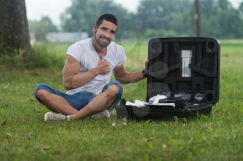 Portrait of Confident Young Engineer Sitting On Grass With Case from Drone and Gives Thumbs Up