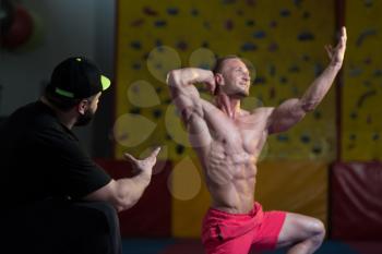 Portrait Of A Man Posing Bodybuilding Poses In Fitness Center and Personal Trainer Corrects Him
