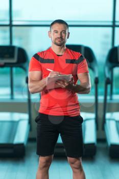 Handsome Personal Trainer With A Clipboard In The Gym And Flexing Muscles - Muscular Athletic Bodybuilder Fitness Model