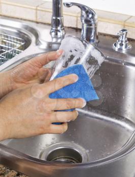 Vertical photo of female hands cleaning a drinking glass with soapy water and a sponge with kitchen sink in background