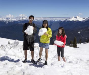 Horizontal Shot of a young man, mature mother and her daughter each holding large snowballs with Snowcap Mountains, trees and blue sky in background
