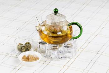 Horizontal photo of glass tea pot, holder and tea balls, sugar in bowl, with White Striped Table Cloth in background