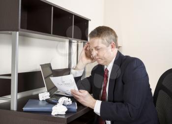 Mature man holding pen in hand against head with computer, calculator, and papers on desk