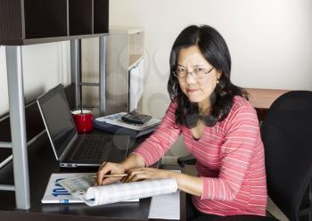 Mature Asian woman  angry doing income taxes with tax form booklet, calculator, coffee cup and computer on desk