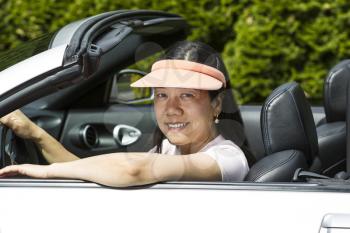 Horizontal photo of a mature women sitting in a convertible car with arm on top of door while wearing a short sleeve shirt and sun visor, outdoors on a lovely day
