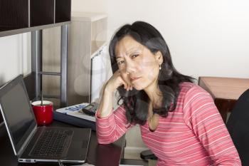 Mature Asian woman tired of doing income taxes with tax form booklet, calculator, coffee cup and computer on desk