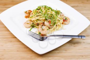 Closeup horizontal photo of Pasta dish with large shrimp, basil, parsley, stainless steel fork on white square plate with natural bamboo wood in background