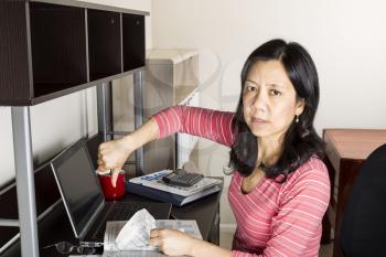 Mature Asian woman giving thumbs down with income tax tables booklet, computer, coffee cup and glasses on desk
