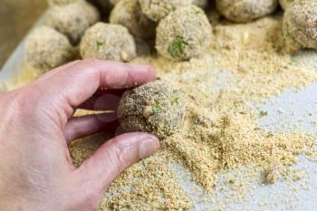 Horizontal photo of male hand holding bread coated raw meatball with bread crumbs and finished meatballs in background