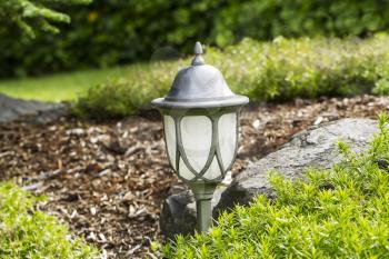 Photo of electric outdoor lamp with post in flower bed with rocks and green in background 
