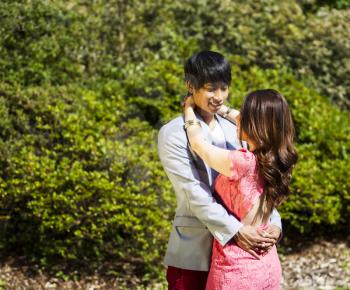 Horizontal photo of young adult couple dressed in formal attire holding each other with green bushes in background 