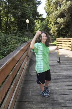 Vertical photo of young girl, looking at small trout, while fishing off of wooden bridge for trout with walk path and trees in background  