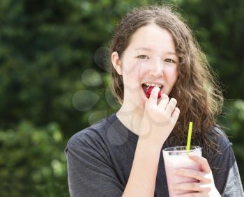 Photo of young girl eating whole strawberry while holding milkshake in other hand with blurred out bright green trees in background 