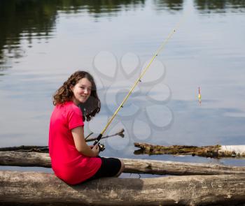 Photo of young girl relaxing, by sitting on logs, before her next cast with lake and trees in background 
