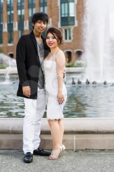 Vertical photo of young adult couple, looking forward, holding each other with water fountain, geese and brick building in background 