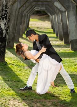 Vertical photo of young adult couple dancing outdoors together underneath a structured column wall in background 