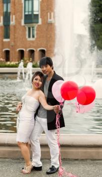 Vertical photo of young adult couple holding several balloons in front of them with water fountain, flowers, trees and brick building in background 