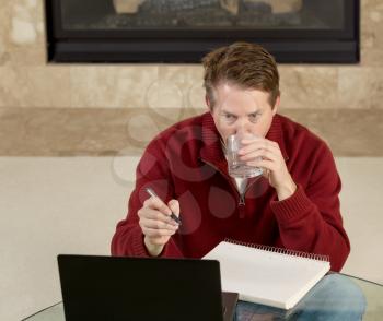 Photo of mature man, drinking water while sitting down at glass table, working from home, looking at computer screen with fireplace in background 