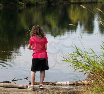 
Photo of young girl, back to camera, scanning lake before casting with lake and trees in background and tall weeds to the right of her 
