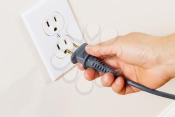 Horizontal photo of female hand inserting power cord receptacle into electric wall outlet 