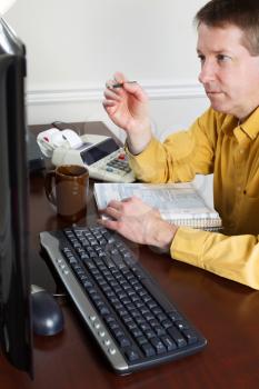Vertical photo of mature man, looking at monitor, working on his taxes with tax booklet and office equipment in background 