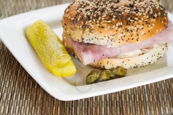 Horizontal photo of a ham sandwich with fresh sliced pickle and capers on white plate with bamboo mat underneath