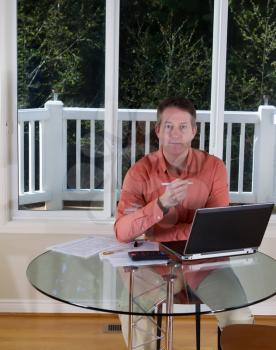 Vertical photo of mature man holding pen in hand, looking forward, while working at home with laptop, calculator and papers on top of table and large windows in background