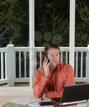 Vertical photo of mature man on cell phone while working at home with laptop, calculator and papers on top of table and large windows in background