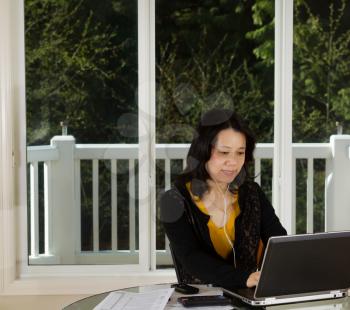Photo of mature woman listening to music while working at home with laptop, cell phone, calculator and papers on top of table and large windows in background