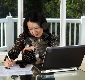 Photo of mature holding her pet while working at home with laptop, calculator, cell phone and papers on top of table and large windows in background