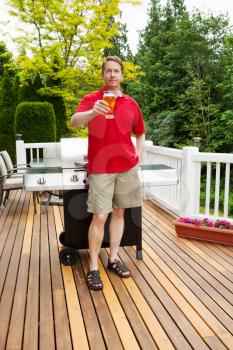 Vertical photo of mature man holding glass of beer with barbecue cooker and seasonal trees in background 