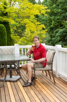 Vertical photo of mature man looking forward and smiling while holding his glass of beer at the table on open cedar patio with seasonal trees in full bloom in background 