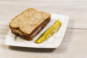 Horizontal photo of ham sandwich made with fresh whole wheat and single large sliced pickle on white plate with faded wood underneath