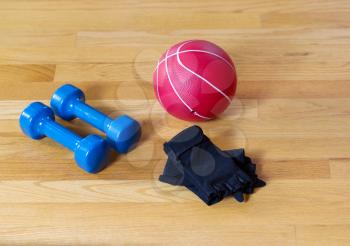 Workout gloves, dumbbells and weight ball lying on wooden gym floor 