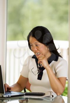 Vertical image of mature woman biting her eye glasses while working from home with blurred out daylight coming in from window in background
