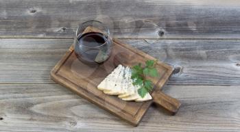Horizontal top view photo of sliced blue cheese, parsley on the side, on a traditional wooden server with a glass of red wine resting on rustic wood