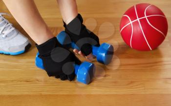 Horizontal image of female hands wearing workout gloves while lifting small weights off of wooden gym floor with red ball and shoe in background