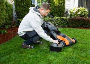 Photo of mature man putting in battery in electric lawnmower on freshly cut plush green grass with home in background 