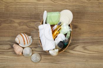 Overhead view of bath and shower accessories placed in basket on rustic wooden boards. Items include hand towel, scrub brush, lotion, soap, soaking salts, perfume, and sea shells.  