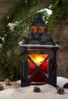 Vertical front view of an old metal lantern, glowing red candle inside, with falling snow and an evergreen tree branch plus rustic wood in background 