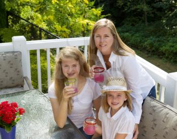 Top front view of happy mother and daughters, looking forward, drinking grapefruit juice while outdoors on patio with woods in background 